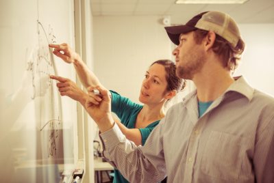 A woman and a man stand facing a white board. The woman points to the board while the man holds a marker.