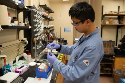 A research pours a solution from one vial into another. He wears safety glasses, a blue lab coat, and blue gloves. He is next to a sink and other lab supplies.