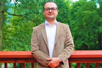 Luis Escobar stands against a wooden deck railing with trees in the background