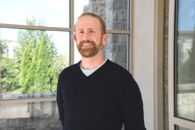 A man wearing a v-neck sweater poses for the camera outside a lab building.