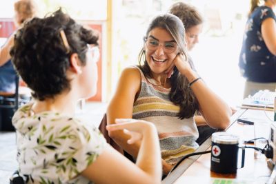 Two women laugh and talk sitting at a table strewn with mugs of coffee, notebooks, and pens.