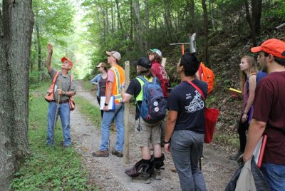 : A group of people holding tools stand on a forested trail, while one person stands near a tree, gesturing towards it. 