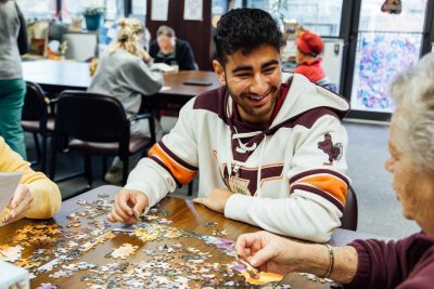 Omar Almasri works on a jigsaw puzzle with an older female participant in a maroon jacket at the Engagement Center for Creative Aging, smiling as they chat.
