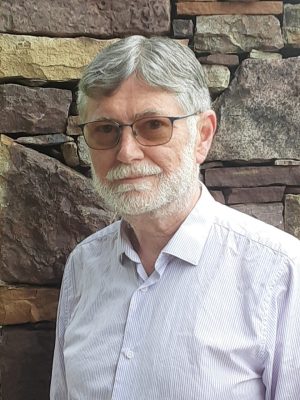 A man with gray hair and a beard is wearing a pin-stripe light-colored dress shirt and glasses, smiling, in front of a stone wall.
