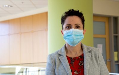 Michelle Olsen, wearing a COVID mask, red blouse, and gray blazer, poses for a photograph inside the Life Sciences 1 building on campus.