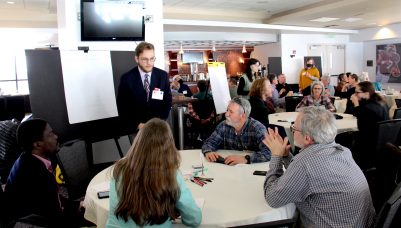 A group of people gathered around a table, with one individual standing to take notes at a notepad on an easel.