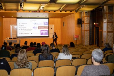 This image shows Linsey Marr addressing  a crowd in an auditorium with a slide presentation in the background.