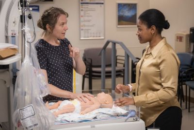 Two women talk over a mannequin baby in a hospital bed